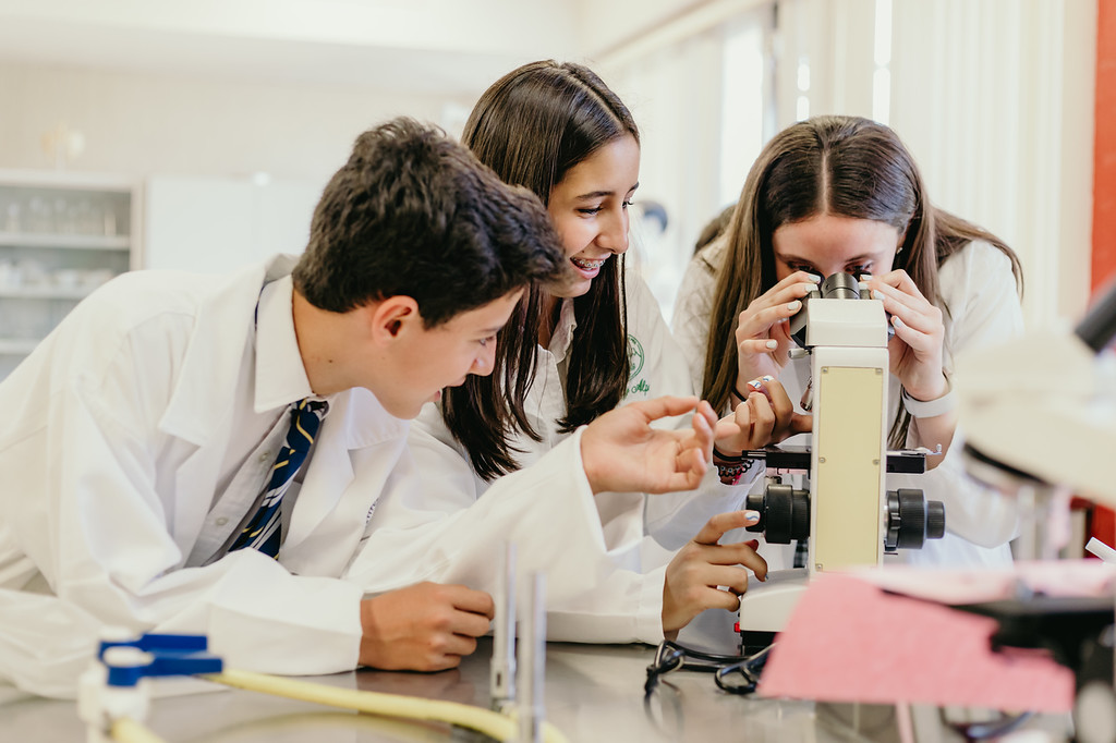 Tres estudiantes 2 mujeres y un hombre vestidos con batas de laboratorio usando un microscopio de las instalaciones de cumbres torreón